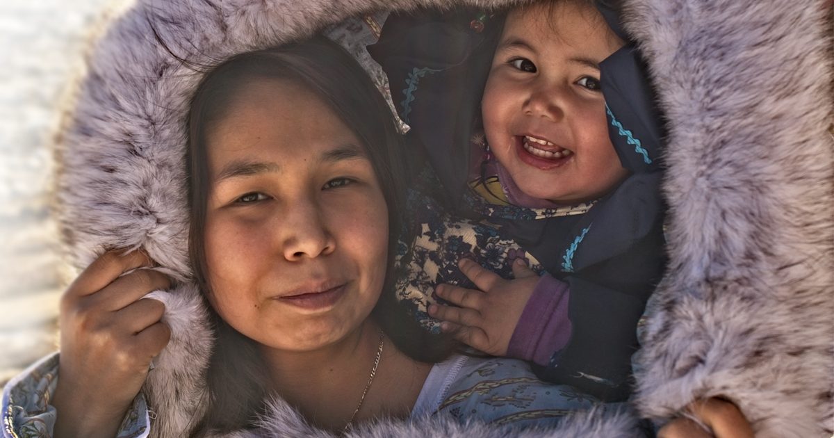 The faces of an Inuit mother and toddler are encircled by the fur of her parka hood.