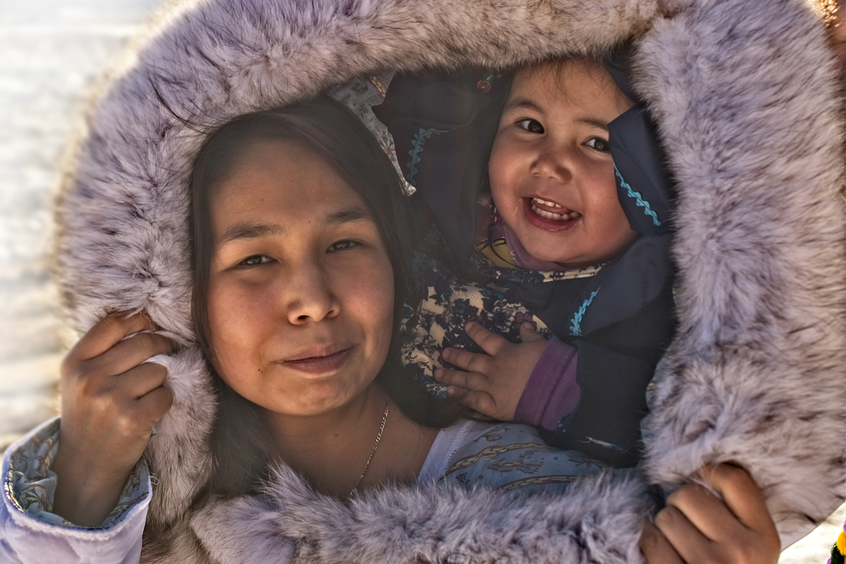 The faces of an Inuit mother and toddler are encircled by the fur of her parka hood.