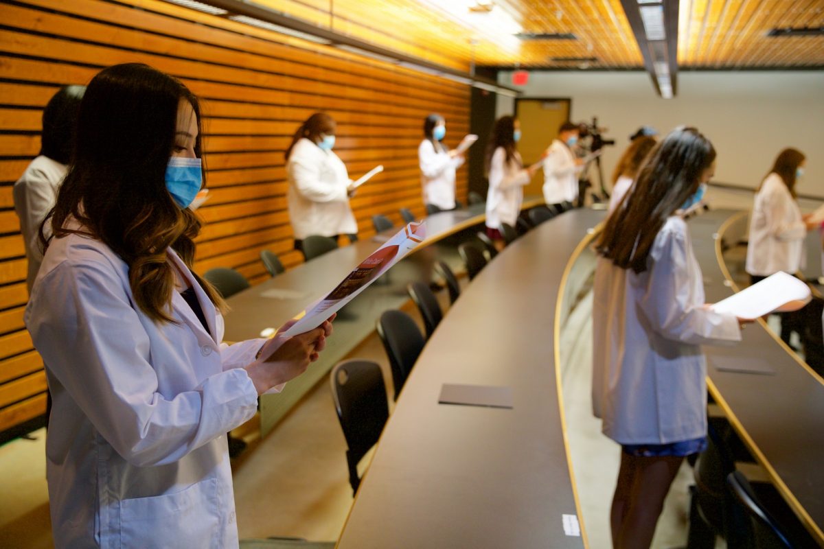 Students stand while wearing white coats and each read off of papers.