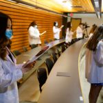 Students stand while wearing white coats and each read off of papers.
