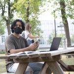 A person with dark curly hair wearing a black face mask sits at a picnic table on campus. They are holding a cell phone and have an open book and laptop in front of them. They are looking off to the left of the camera.