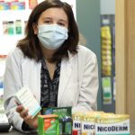 A woman wearing a white coat and surgical mask stands behind a pharmacy counter with smoking-cessation products in front of her.