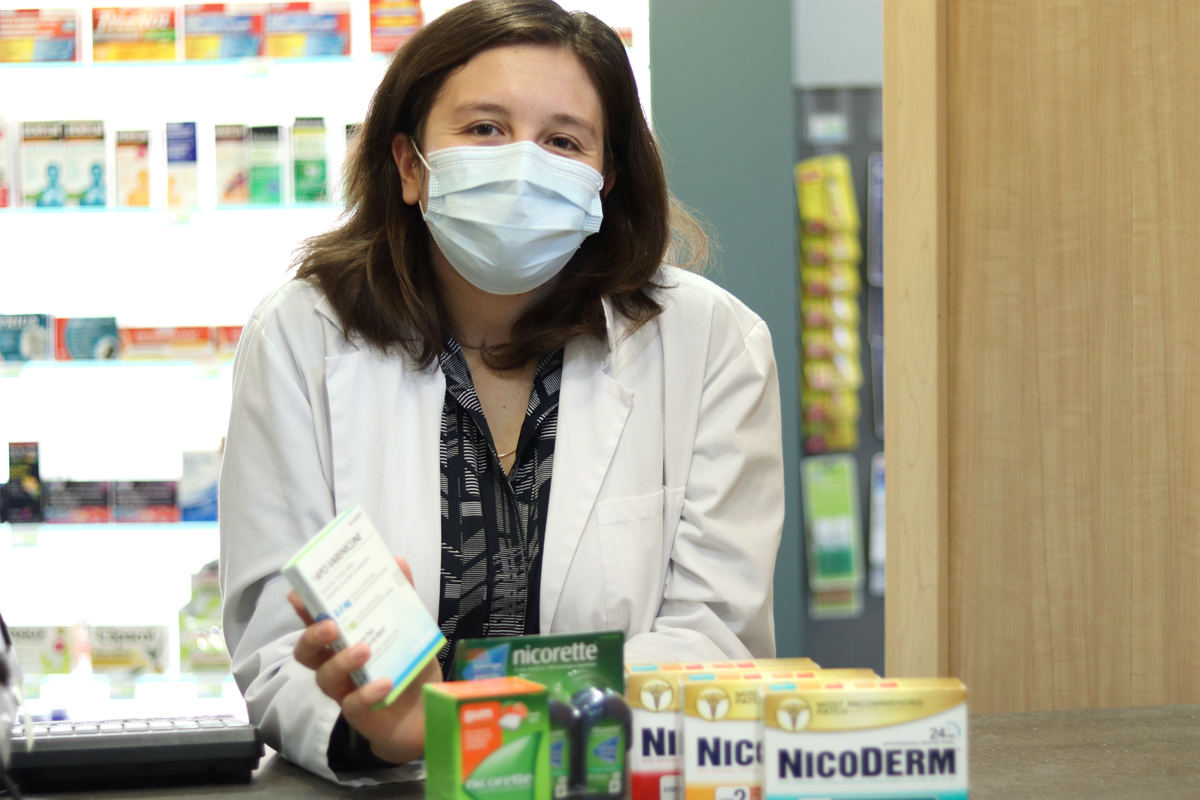 A woman wearing a white coat and surgical mask stands behind a pharmacy counter with smoking-cessation products in front of her.