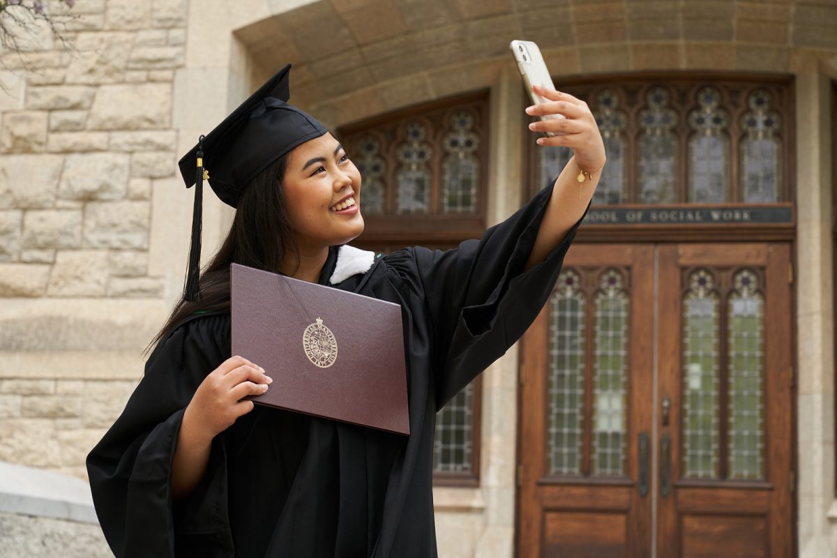 A graduating student poses for a selfie with her UM Diploma. She stands in front of a brick building with ornate doors wearing a black cap and gown. She holds a phone in her left hand and smiles widely.