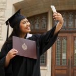 A graduating student poses for a selfie with her UM Diploma. She stands in front of a brick building with ornate doors wearing a black cap and gown. She holds a phone in her left hand and smiles widely.