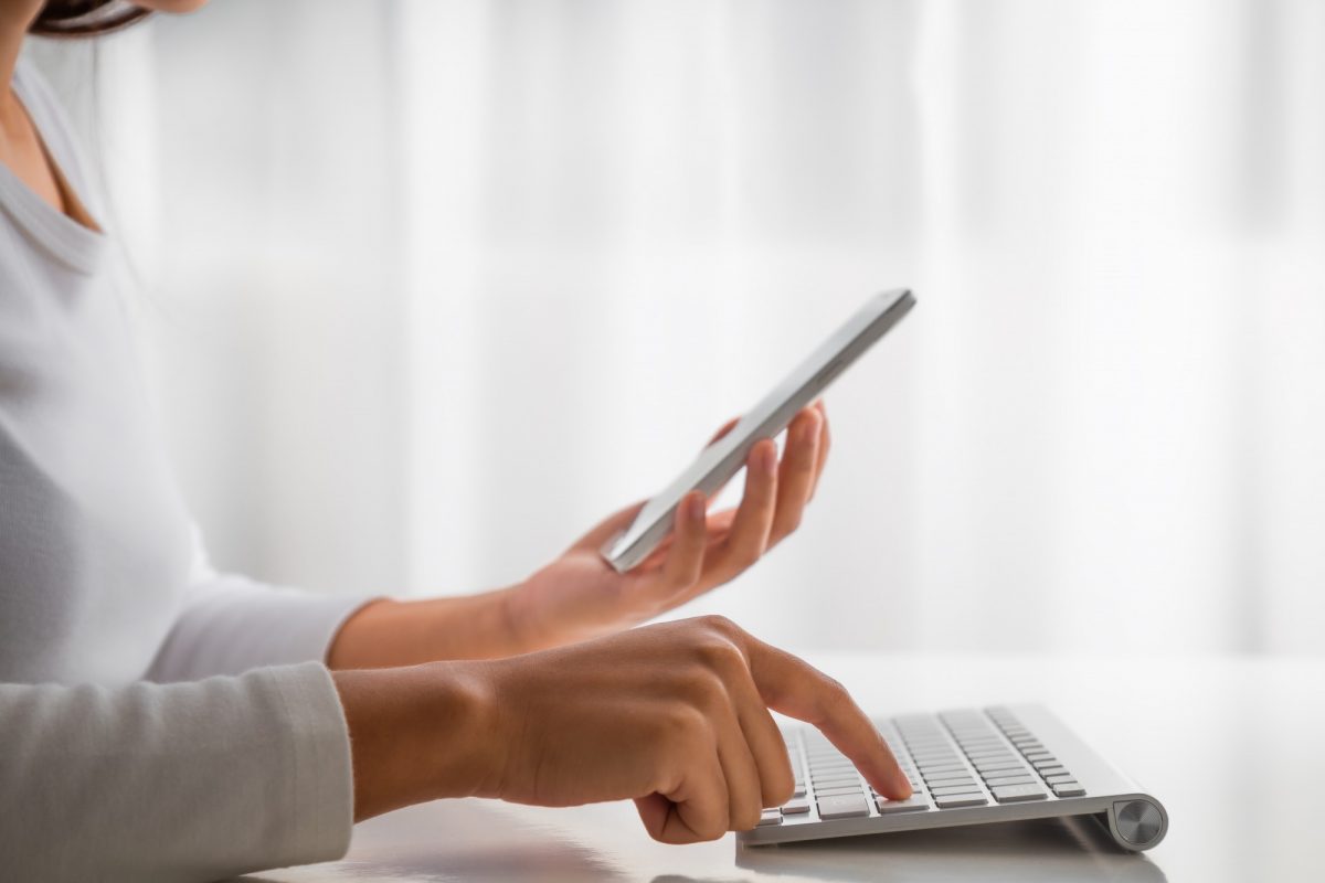 Hands typing on keyboard and looking at mobile phone.