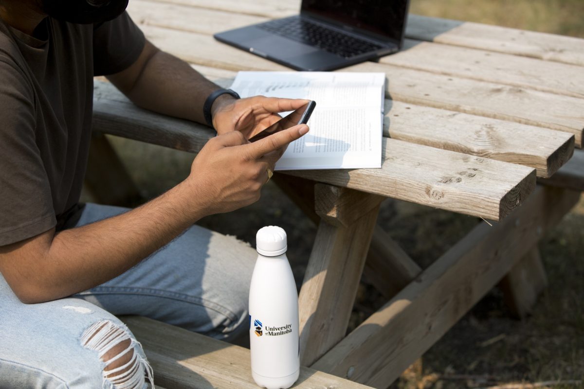 A person sits at a picnic table and scrolls on a smart phone. A laptop and an open text book sit on the table behind them, and a white water bottle with the UM logo on it sits on the bench in front of them.