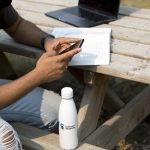 A person sits at a picnic table and scrolls on a smart phone. A laptop and an open text book sit on the table behind them, and a white water bottle with the UM logo on it sits on the bench in front of them.