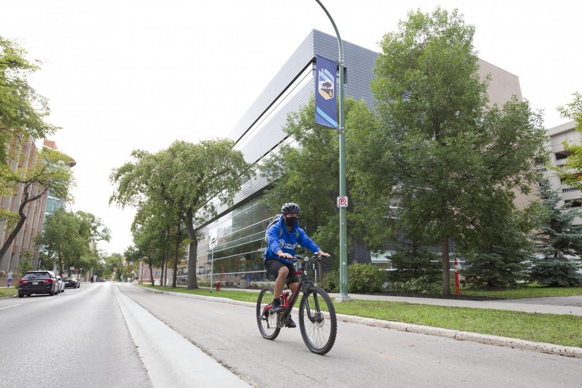 A person wearing a bike helmet, mask and blue UM hoodie rides a bike on the Bannatyne Campus
