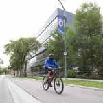A person wearing a bike helmet, mask and blue UM hoodie rides a bike on the Bannatyne Campus