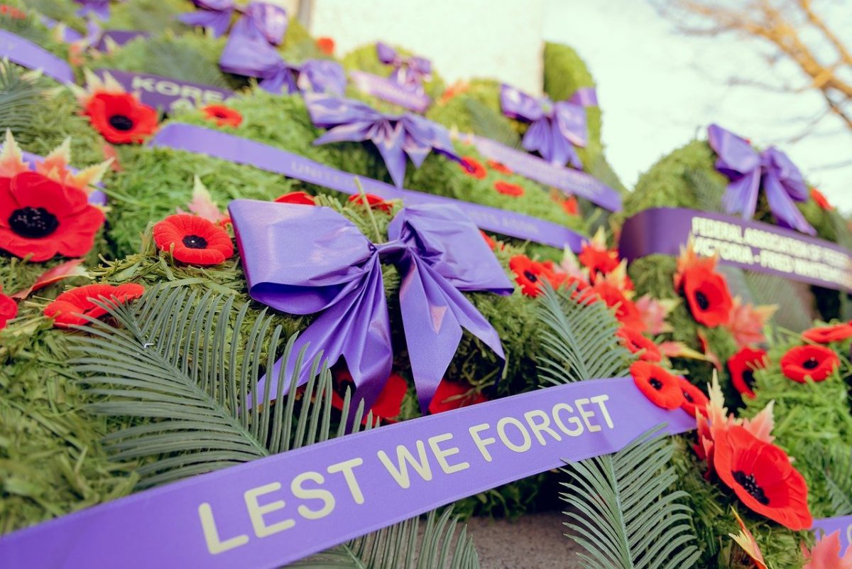 Green wreath with poppies and a purple ribbon. On the ribbon are the words Lest We Forget.