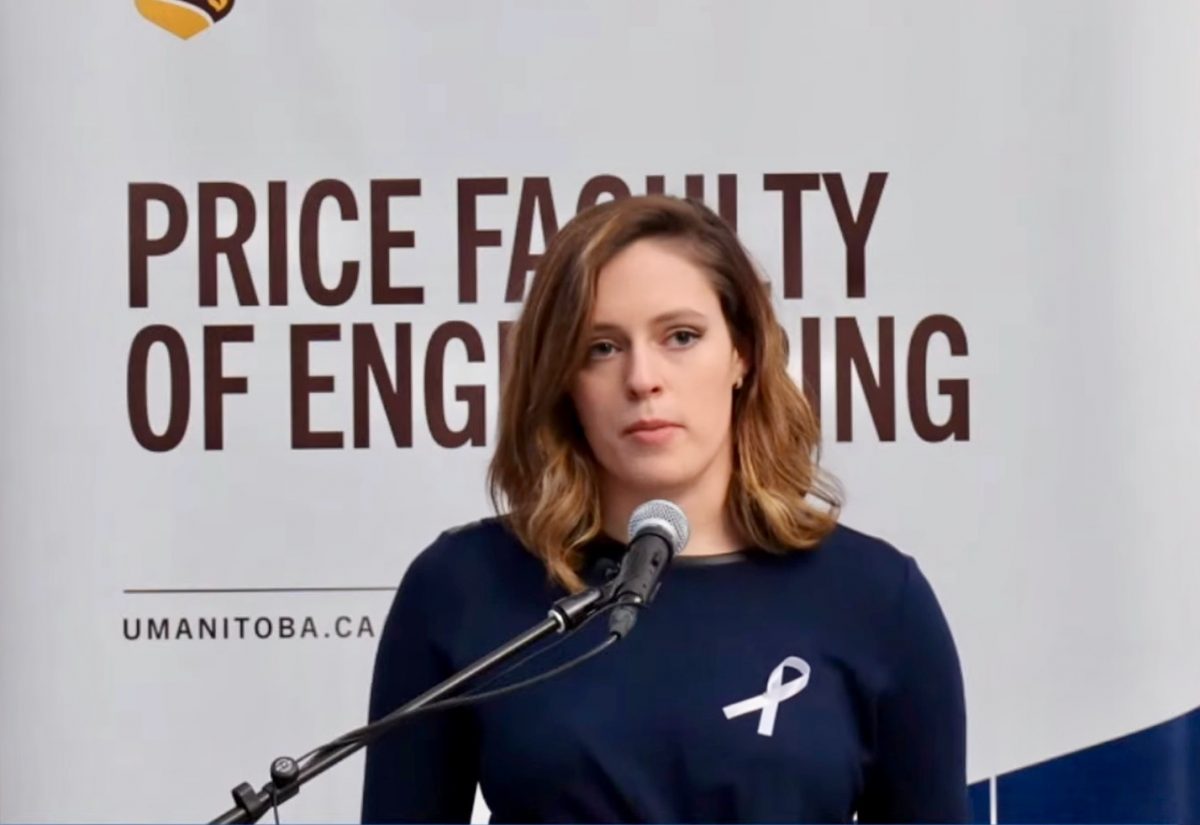 Avery Edwards stands with a netural expression while standing at a podium with a microphone. She is wearing a white ribbon on her right lapel.