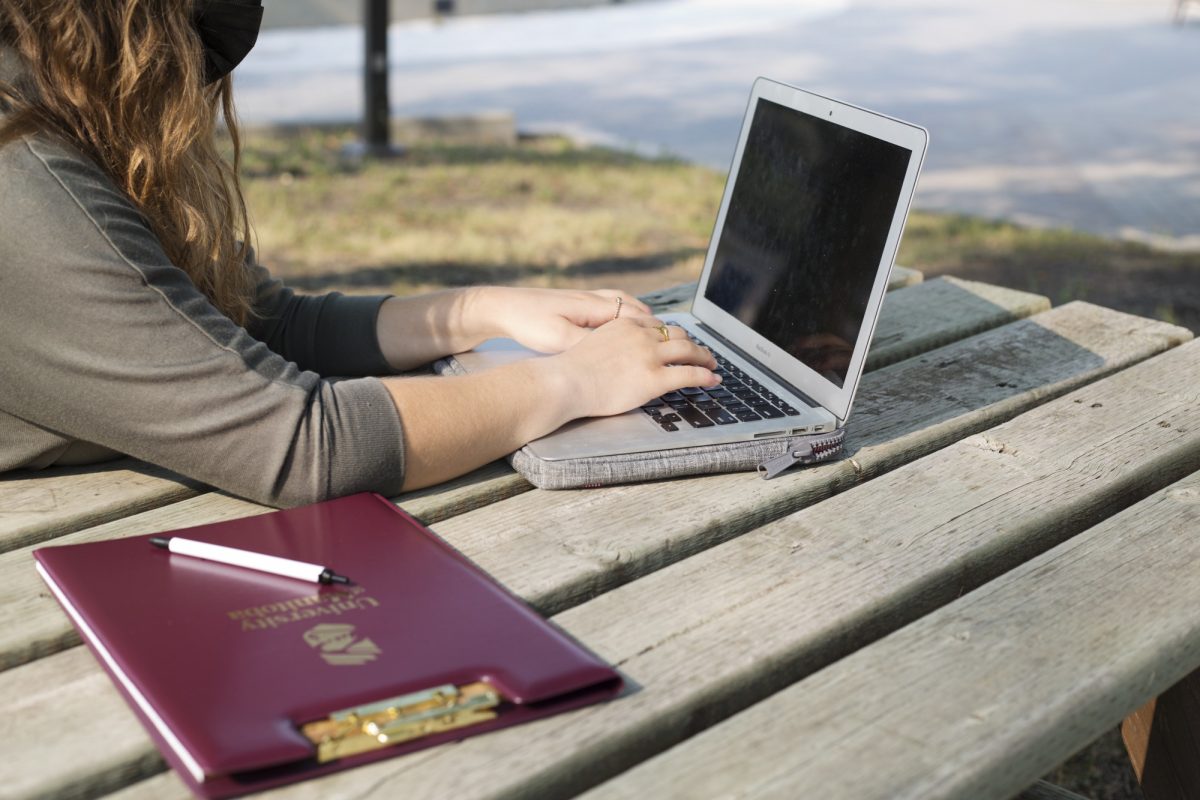 A red UM clipboard sits on a picnic table in front of a person typing on a laptop computer.