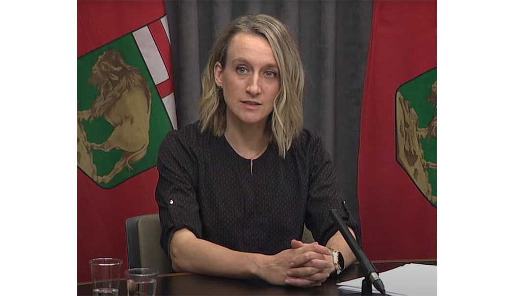 Dr. Joss Reimer at a microphone during a press conference. Manitoba flags in the background.