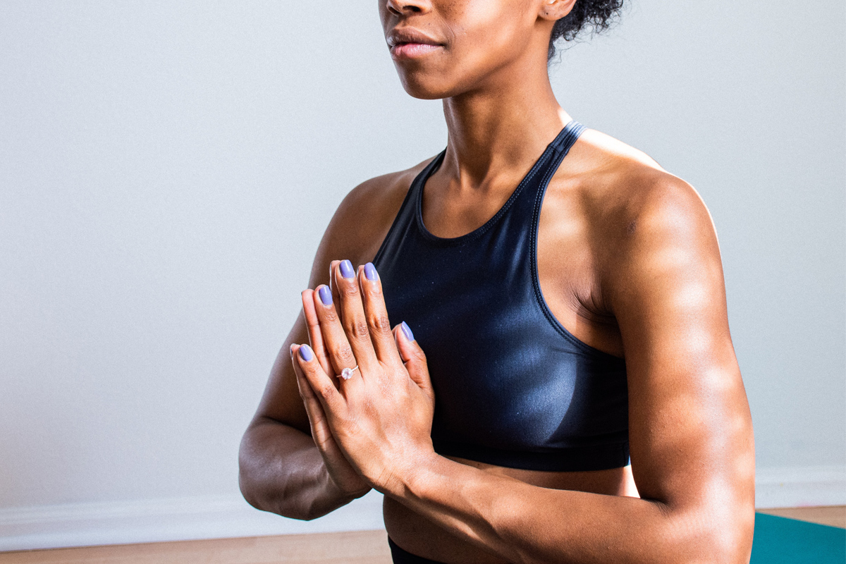 A feminine person sits meditating on a yoga mat with their hands pressed together