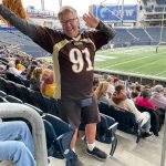 Man in brown and gold sports jersey strikes a dancing pose in the stands of a football stadium