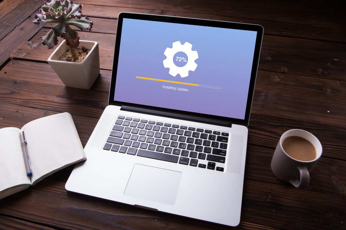 A laptop sits on a desk with a program update loading on its screen. It is surrounded by an open notebook, a cup of coffee and a small potted plant