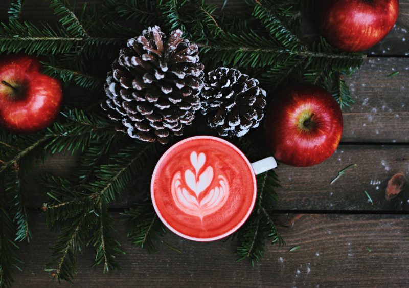 A winter tableau with red apples, pine cones, pine branches and a latte topped with a design in red-tinted foam.