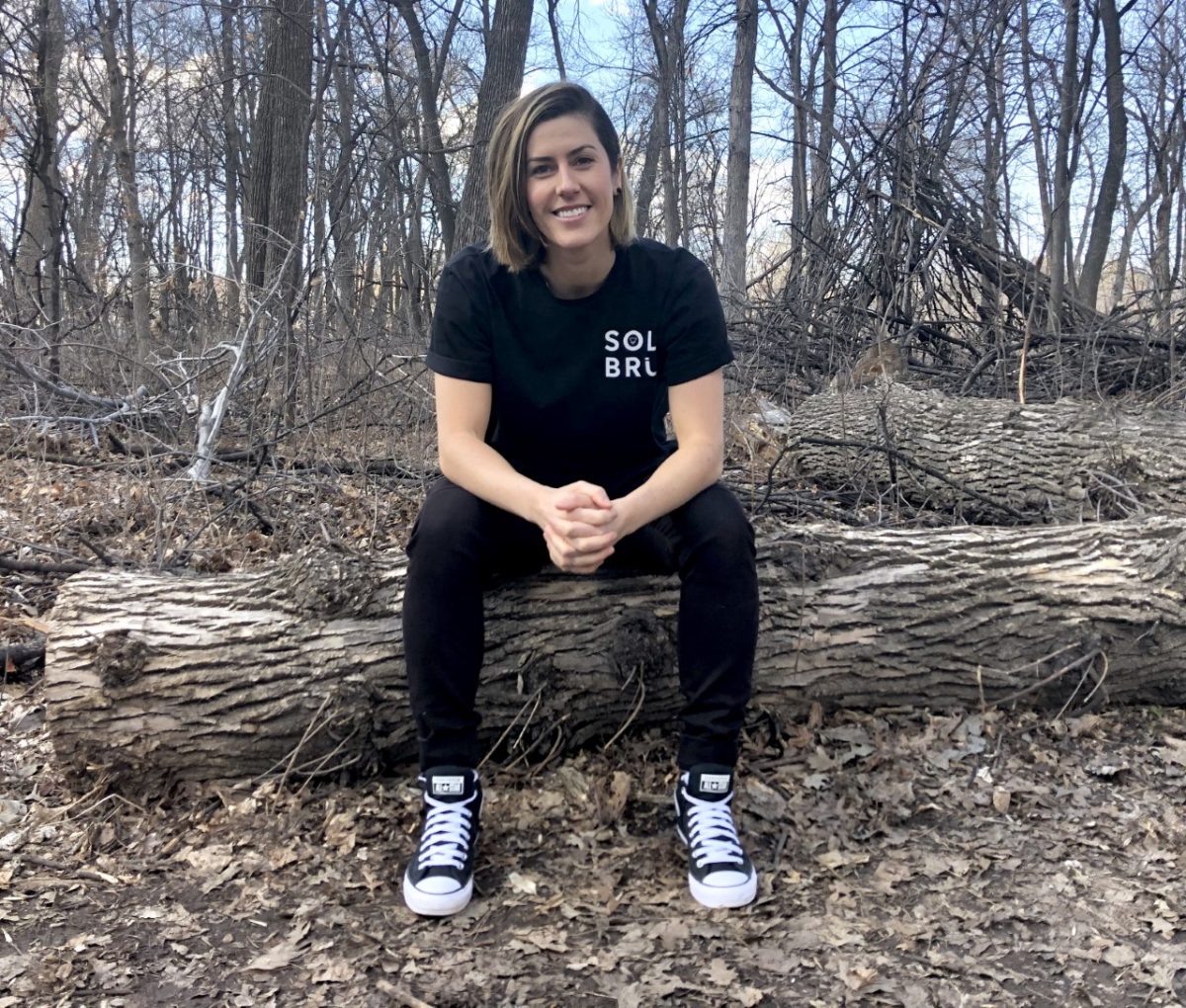 Asper alum Leanne Kisil wearing all black sitting on a log in the forest smiling.