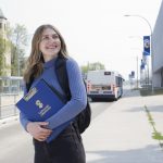 An excited student stands outside on the Fort Garry campus holding a UM clipboard. A bus retreats in the background.