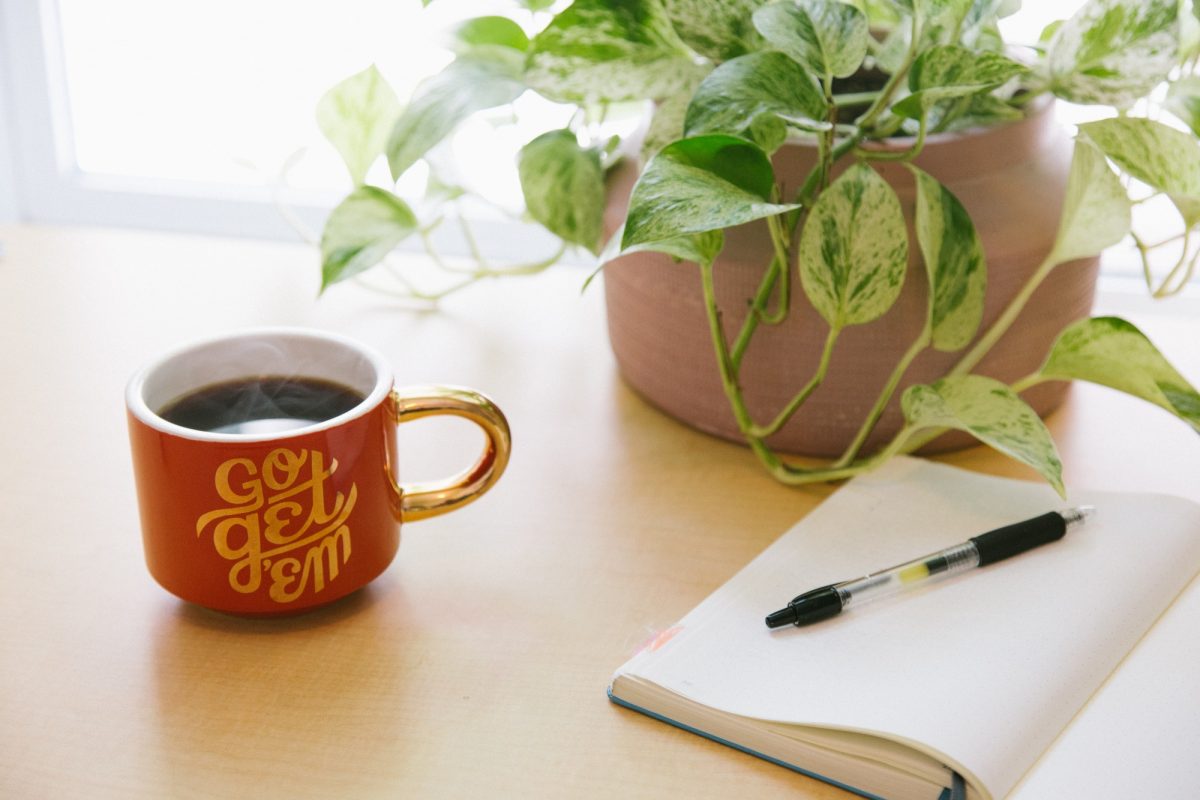 A notebook lies open on a desk beside a mug with an encouraging "go get 'em" message. A plant sits in the background.