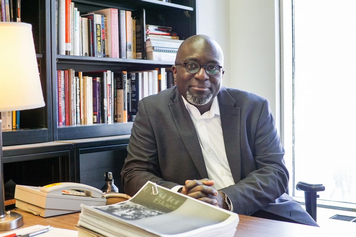 Professor Barrington Walker leaning on his desk with bookshelf behind.