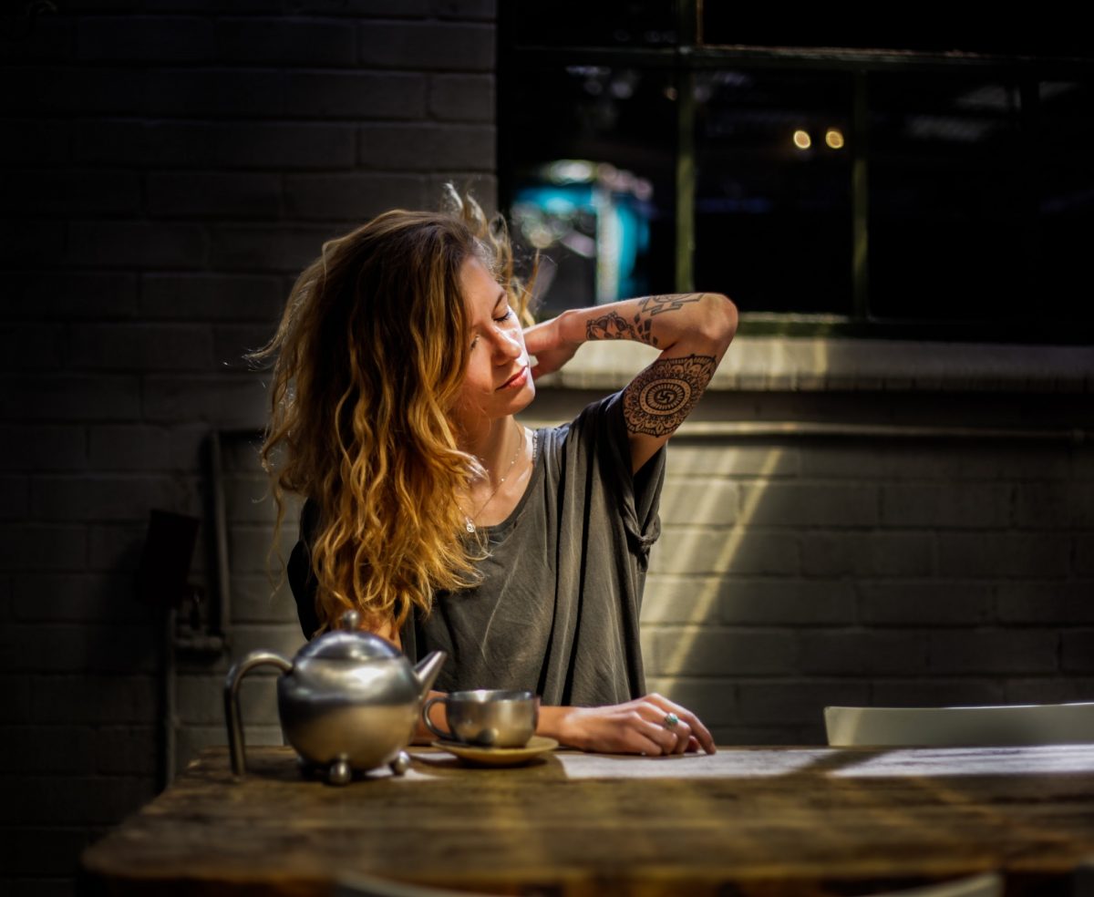 A feminine person with arm tattoos sits at a table, rubbing their tense neck muscles with a peaceful look on their face. A teapot and tea cup sit on the table in front of them.