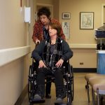A health care worker assists a woman in a wheelchair in a hospital hallway.
