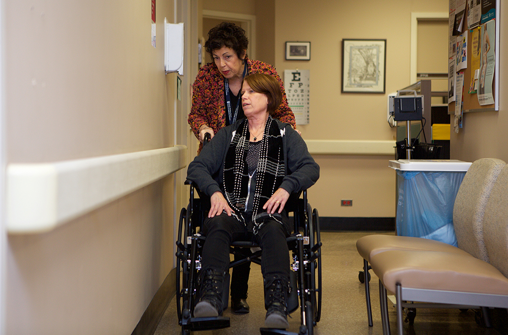 A health care worker assists a woman in a wheelchair in a hospital hallway.