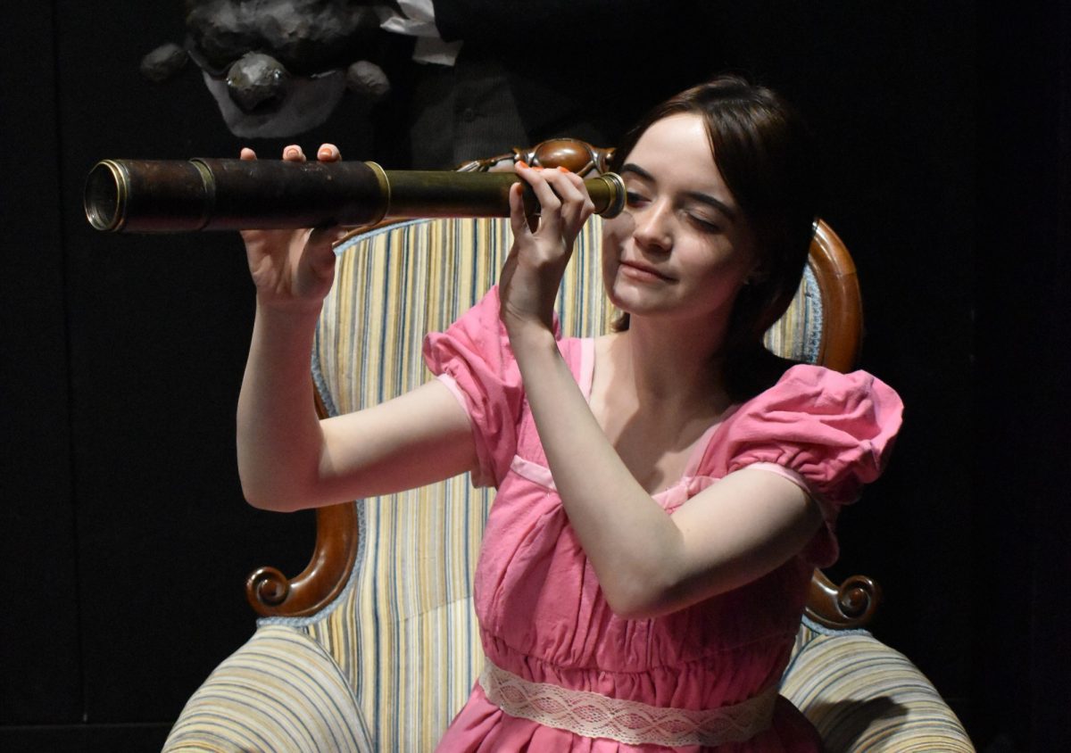 Actress in pink dress sitting on large, plush armchair, looking through an antique telescope.