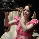 Actress in pink dress sitting on large, plush armchair, looking through an antique telescope.