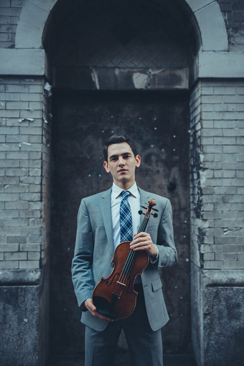 Gregory Lewis stands in a suit and tie with a violin in hand.