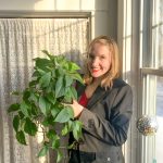 Katherine Hawthorne stands smiling posting for the camera, holding up a medium size green leaf plant.