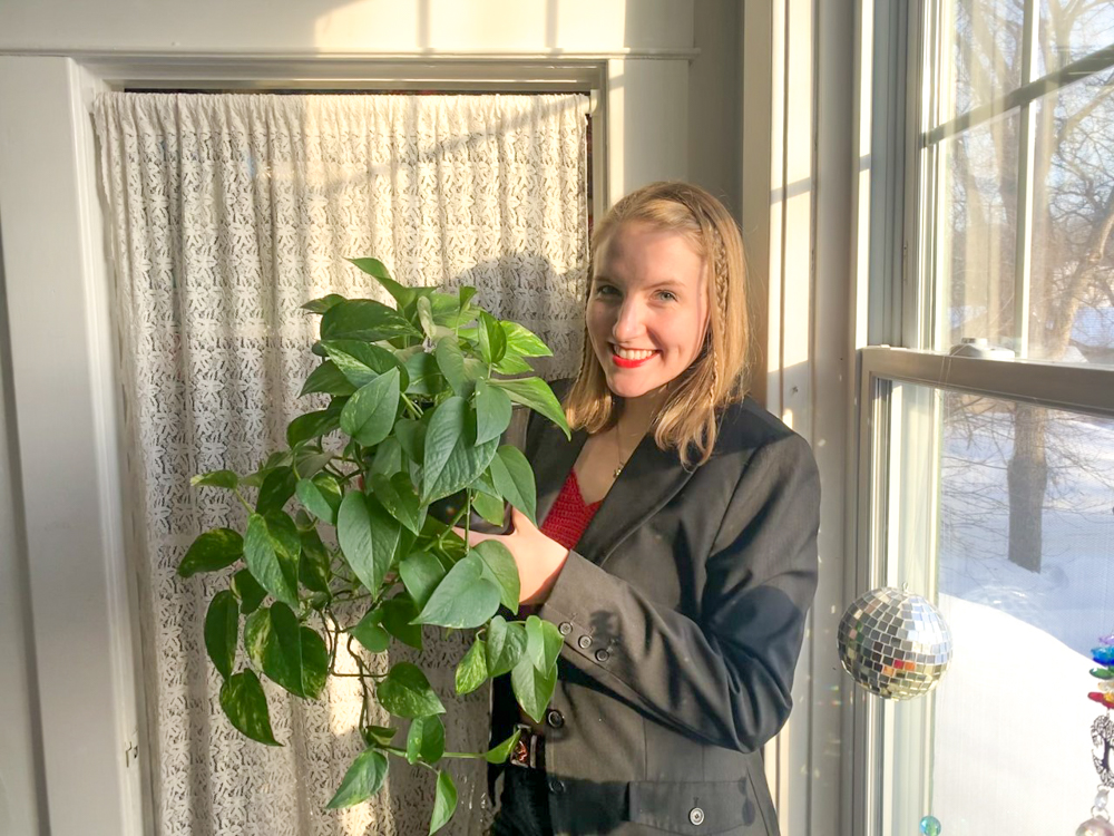 Katherine Hawthorne stands smiling posting for the camera, holding up a medium size green leaf plant.