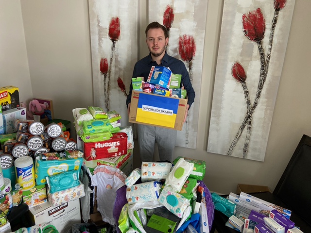 He holds a box with a Ukrainian flag on it and the words "Supplies for Ukraine." The box is filled with medical supplies and he is surrounded by donated items. 