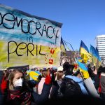 Sign reads "Democracy in Peril." Background of sign is Ukrainian flag and there are two red handprints. People in background of photo holding Ukrainian flags.