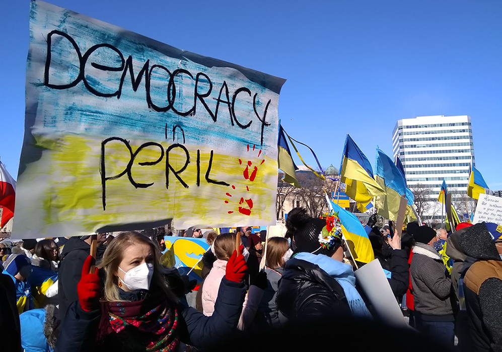 Sign reads "Democracy in Peril." Background of sign is Ukrainian flag and there are two red handprints. People in background of photo holding Ukrainian flags.