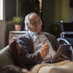 An elderly man looks at a tablet while wearing headphones. A wheelchair is in the background