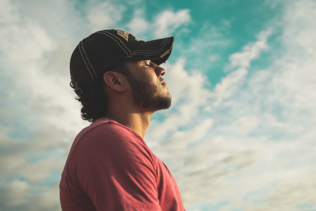A masculine person in a red t-shirt and black ball cap stands against the sky and inhales deeply