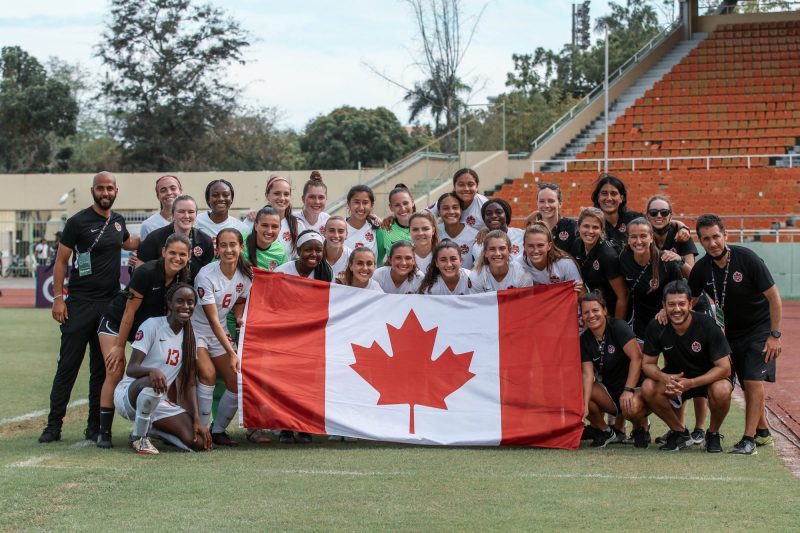 the Canada U-20 (girls) team stands behind a big Canadian flag