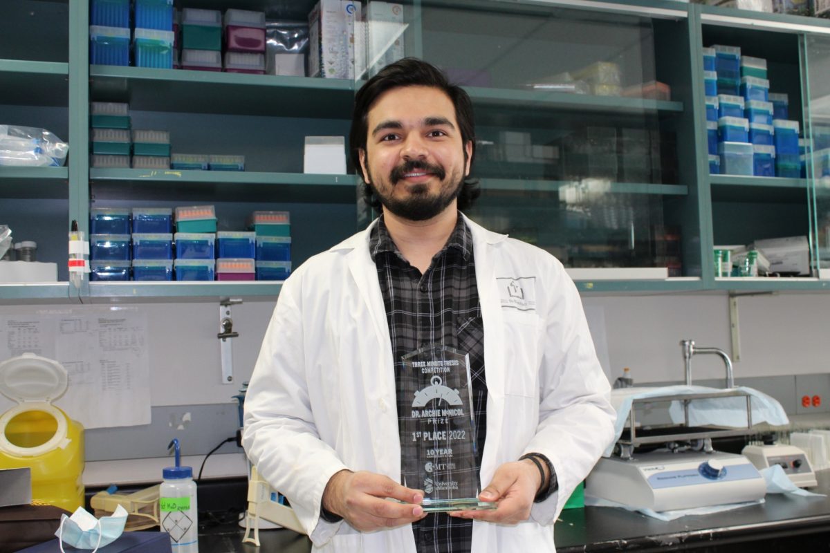 Student wearing a lab coat holds a glass trophy with 3MT logo on it.