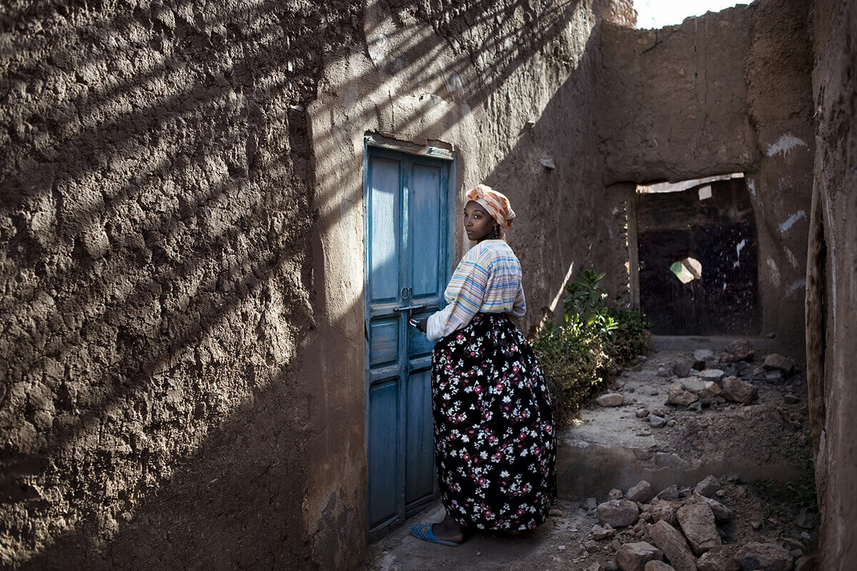 A photo of a woman in a long dress and head wrap stands in front of a blue door in a building with rocks on the ground and a missing portion of a wall.