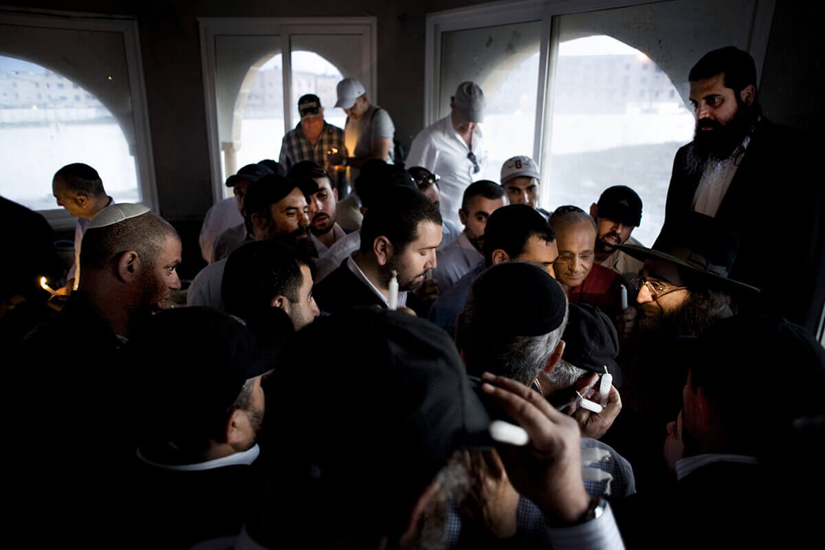 An indoors photo of a group of men gathered around an orthodox rabbi.