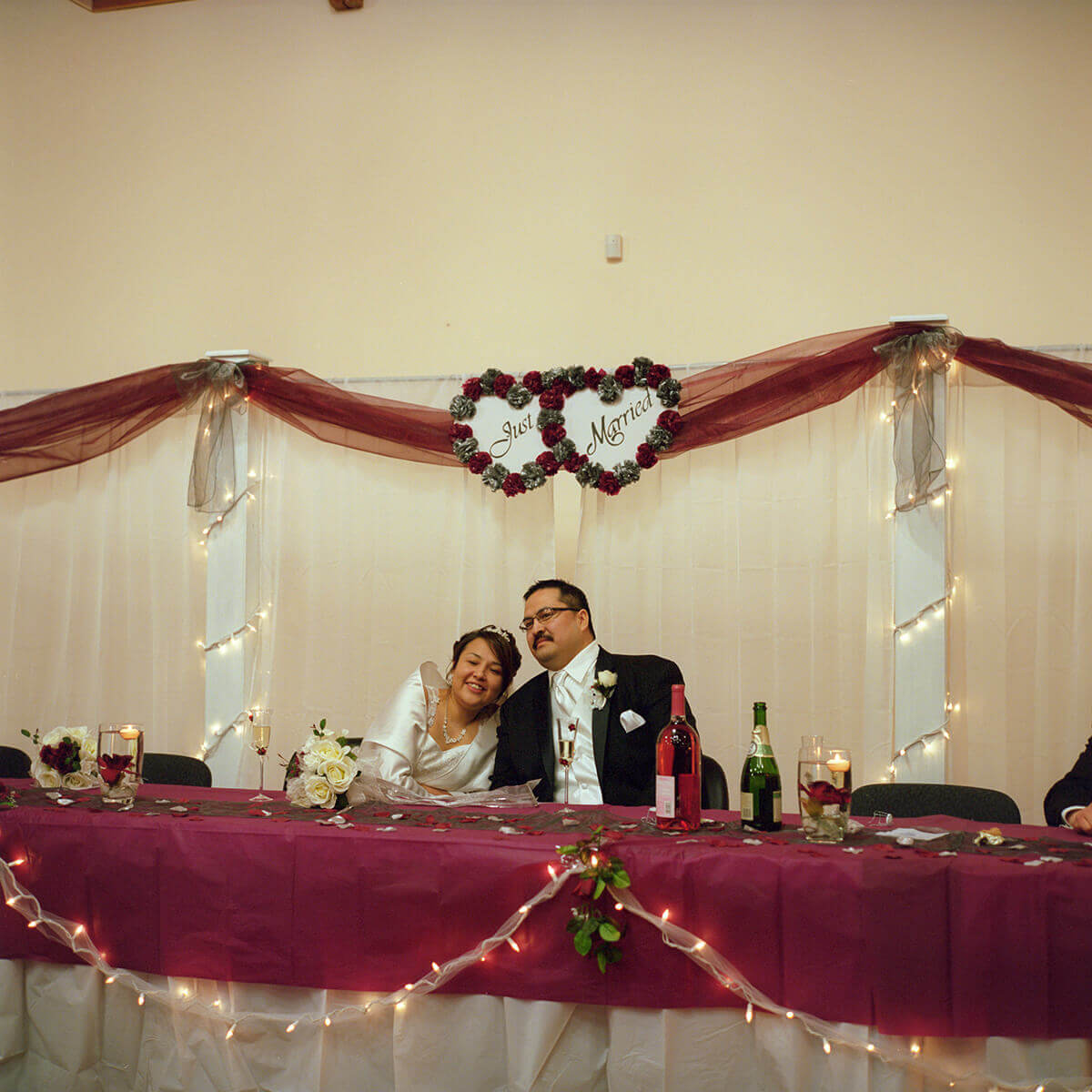 A photo of a couple in wedding clothes sitting at the middle of a table in a wedding venue. Above their heads are two paper heart decorations reading Just Married.