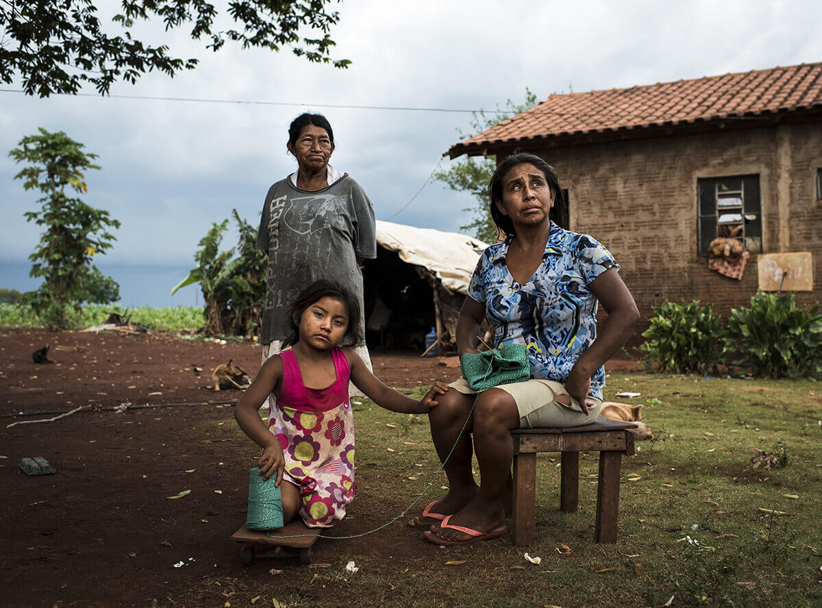 A photo of three generations of Indigenous Brazilian women posing together. The grandmother stands tall behind the daughter, and the mother sits on a chair next to her.