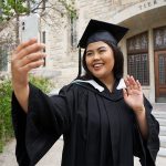 A new grad in cap and gown waves as she takes a selfie
