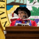 Dr. Mary Jane McCallum stands at a lectern. She holds an eagle feather and wears an honorary Doctor of Laws degree gown and hat.