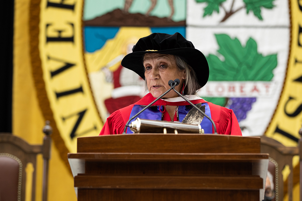 Dr. Mary Jane McCallum stands at a lectern. She holds an eagle feather and wears an honorary Doctor of Laws degree gown and hat.