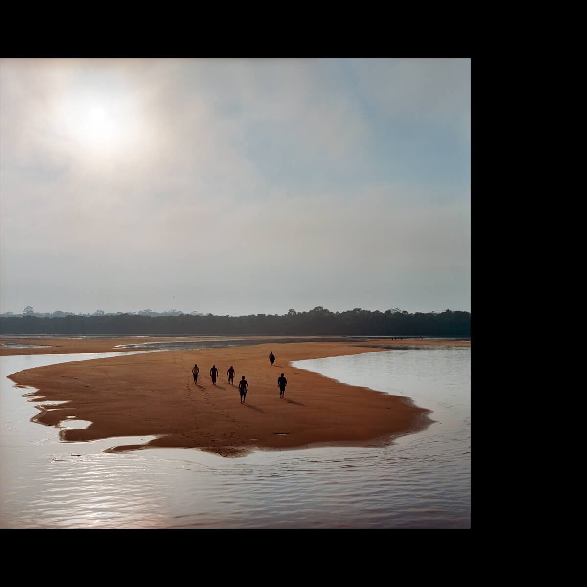A photograph in which a group of people walk on a sandbar underneath a bright, clouded sun. The photo is offset in a field of plain black.