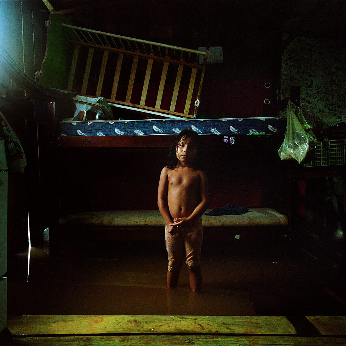 A photo of a girl standing in a bedroom that has been flooded with dark, murky water. He is staring directly at the camera.